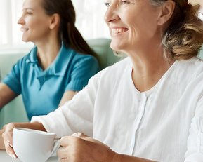 Eine ältere Frau mit grauen Haaren hält eine Tasse in der Hand und lächelt. Neben ihr sitzt eine jüngere Frau in einem blauen Shirt. Auf dem Tisch liegt ein Stück Kuchen und ein roter Stift.