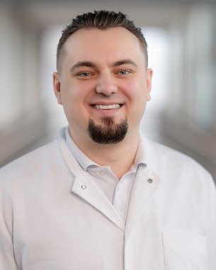 Ein lächelnder Mann in einem weißen Kittel steht in einem modernen Innenraum. Er hat einen kurzen Bart und trägt ein Hemd darunter.