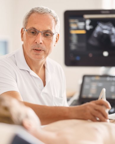 Ein Arzt führt einen Ultraschalluntersuchung bei einem Patienten durch. Der Arzt trägt eine Brille und hat graue Haare. Im Hintergrund ist ein Ultraschallgerät sichtbar, das die Untersuchung unterstützt.