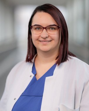 Eine Person mit kurzen, braunen Haaren und Brille trägt einen weißen Kittel über einem blauen Oberteil. Sie steht in einem hellen, modernen Flur.