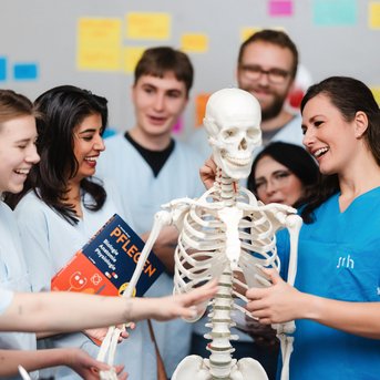 Eine Gruppe von Personen in blauen Arbeitskleidung steht um ein menschliches Skelett und diskutiert, während eine Person ein Buch mit dem Titel "Pflegen" hält.