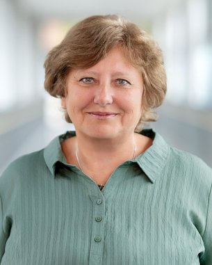 Porträt einer Frau mit kurzen, lockigen Haaren, die ein grünes Hemd trägt. Sie lächelt freundlich in die Kamera.