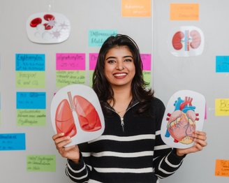 Eine Person hält Modelle von Lunge und Herz in den Händen, während sie vor einer Wand mit bunten Notizen steht.
