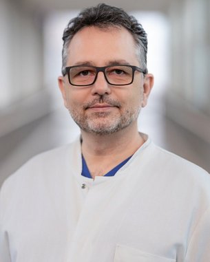 Ein Mann mit Brille und Bart trägt einen weißen Kittel. Er steht in einem hellen Flur und schaut direkt in die Kamera.